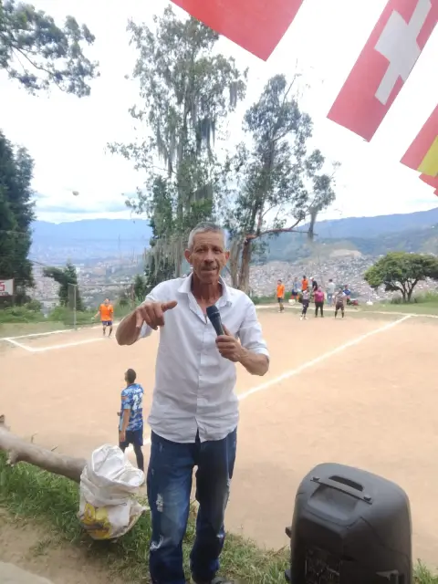 Jaime Ramirez con micrófono junto a una cancha de tierra con banderines y el valle de Medellín al fondo